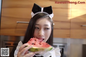 A woman in a maid outfit washing a watermelon in a kitchen sink.