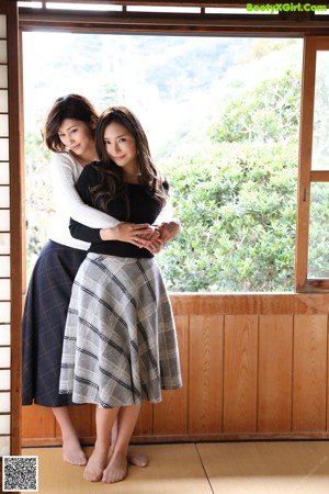 A woman in a black sweater is holding a young woman's breasts.